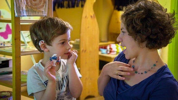 Frau spricht mit einem gehörlosen Kind, welches ein Hörgereät am Kopf trägt