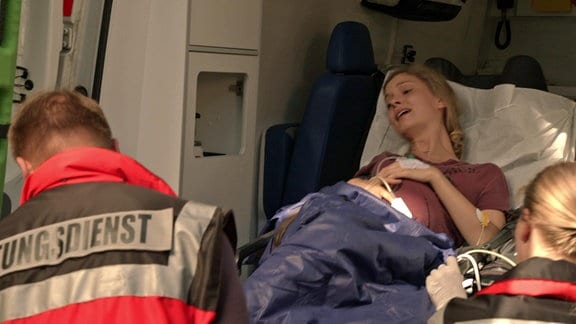 Eine schwangere Frau wird von einer Sanitäterin und einem Sanitäter in einen Krankenwagen geschoben.