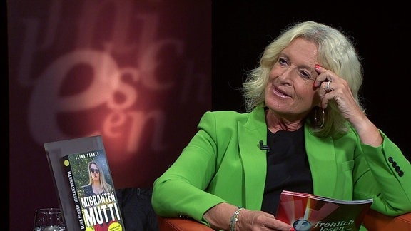 Neben einer sitzenden Frau in grünem Jacket steht ein Buch mit dem Titel „Migranten Mutti“.