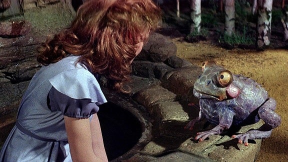 Eine junge Frau sitzt auf dem Rand eines Brunnens und blickt einen Frosch an, der ebenfalls auf dem Rand sitzt.