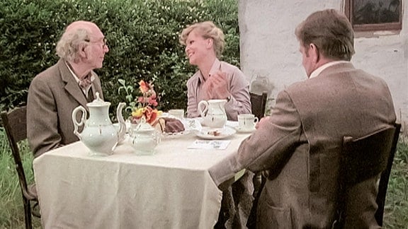 Frau und zwei Männer sitzen bei Kaffee und Kuchen an einem Tisch in einem Garten
