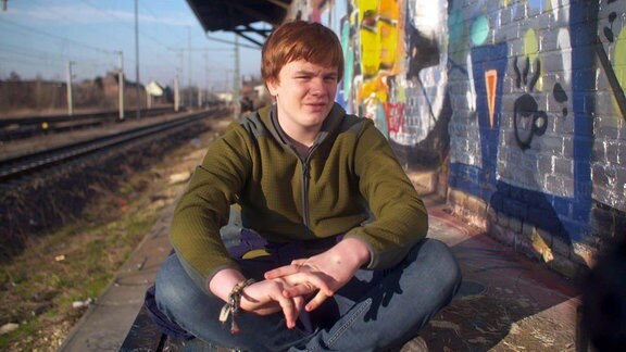 Ein junger Mann sitzt mit verschränkten Beinen auf einer Rampe neben einer mit Graffiti besprühten Wand eines Bahnhofgebäudes und eines Gleisbettes.