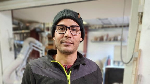 Porträt eines Mann mit Brille und Mütze in einer Werkstatt.