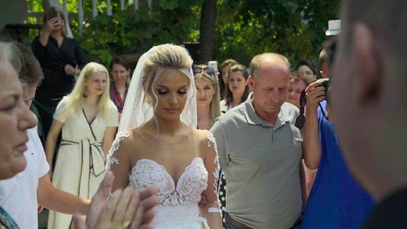 Eine junge Braut mit Schleier und weißem Kleid umringt von Gästen.