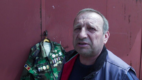 Ein älterer Mann steht vor einer rostroten Gebäude, an dem eine Uniformjacke mit vielen Orden hängt.