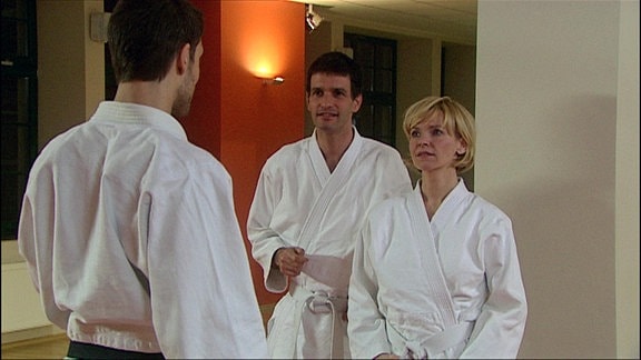 Zwei Männer und eine Frau stehen sich in Judokleidung gegenüber.