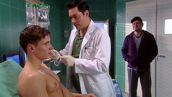 Ein junger Mann sitzt in einem Krankenbett und wird von einem Arzt untersucht. Im Hintergrund steht ein lächelnder Mann mit Basecap in der Tür.