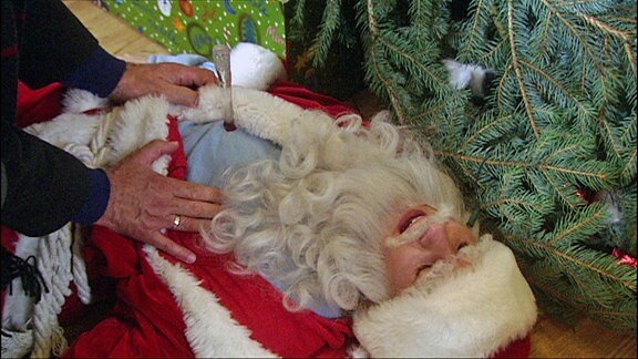Ein am Boden liegender Weihnachtsmann mit zwei fremden Händen auf dem Brustkorb.