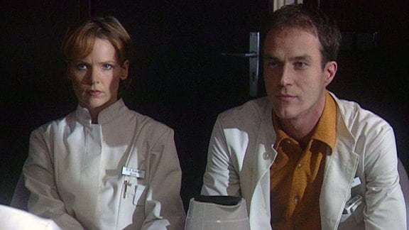 Eine Frau und ein Mann in weißen Kitteln sitzen vor einem Schreibtisch.