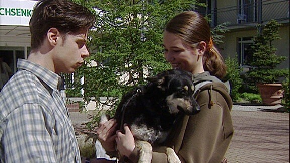 Eine junge Frau mit einem Hund auf dem Arm und ein junger Mann stehen auf einer Straße.