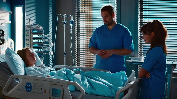 Ein Mann und eine Frau in blauer Kleidung stehen am Krankenhausbett einer Frau.