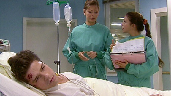 Ein junger Mann liegt mit einer Kopfverletzung im Bett, an dem zwei Krankenschwestern stehen.