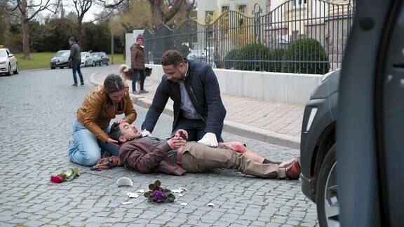 Ein Mann liegt verletzt auf einer Straße. Neben ihm knien eine Frau und ein Mann mit weißen Handschuhen. Ein zerschlagener Blumentopf und eine Rose liegen auf dem Boden daneben.