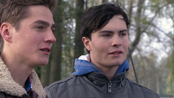 Zwei junge Männer stehen nebeneinander in einem Waldstück.