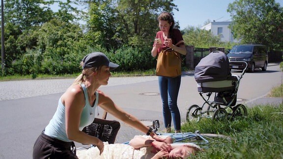 Eine am Boden liegende und eine neben ihr kniende Frau an einem Straßenrand. Hinter einem im Gras liegenden Fahrrad steht eine Frau neben einem Kinderwagen und holt ihr Mobiltelefon aus der Tasche.