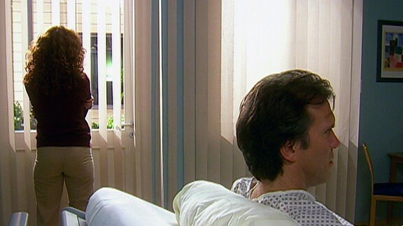 Ein Mann liegt in einem Krankenhausbett. Dahinter, abgewandt, steht eine Frau mit verschränkten Armen und schaut aus dem Fenster.