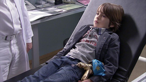 Ein Junge liegt bei einem Arzt auf der Liege. Unter seinem Arm klemmt eine goldbekleidete Puppe mit blauem Sternenumhang.