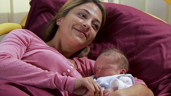 Eine lächelnde Frau liegt mit einem Baby im Arm auf einem Kissen.