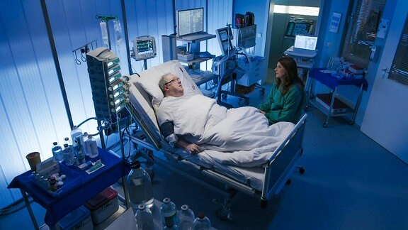Blick in ein Krankenhauszimmer mit vielen Gerätschaften und einer Frau, die am Bett eines Mannes sitzt.