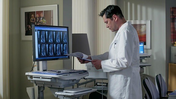 Ein Arzt in weißem Kittel steht vor einem Monitor mit Röntgenbildern und blättert in Papieren. 