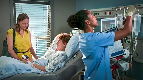 liegender Patient überreicht einer jungen Frau ein Etui, während eine Krankenschwester eine Infusion vorbereitet