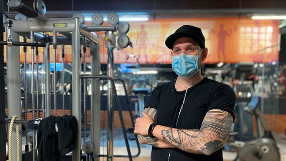 Ein junger Mann mit Maske steht in einem Fitnessraum mit Sportgeräten. 