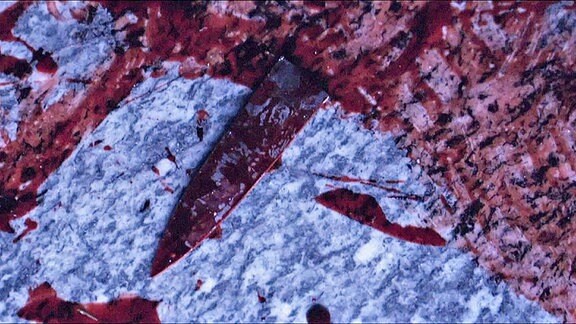 Ein Stück einer blutigen Messerklinge auf einem blutverschmierten Boden.