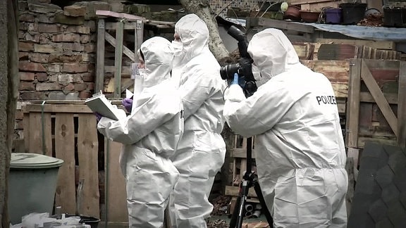 Drei Polizisten in weißen Schutzanzügen und Masken bei der Spurensicherung mit Fotoapperat und Notizblock an einem verfallenen Gebäude.