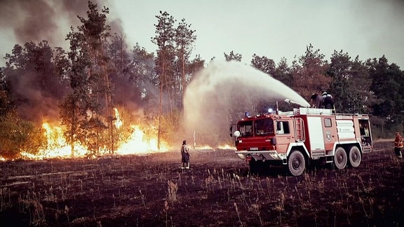 Ein Feuerwehrfahrzeug und zwei Männer löschen einen Waldbrand.