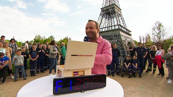 Ein Mann mit Mikrofon steht an einer Kiste vor einem kleinen Eifelturm und  Zuschauern. 