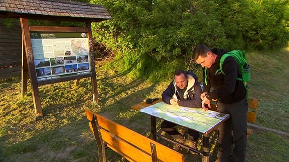 Zwei Männer mit Rucksack schauen an einem Rastplatz auf eine Karte, die auf einem Tisch liegt.  
