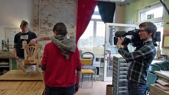 Drei Menschen betrachten einen Schaukelstuhl und werden dabei von einem Kameramann gefilmt.