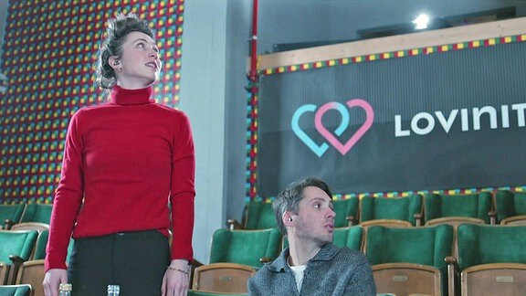 Eine junge Frau und ein junger Mann in einem Hörsaal oder Kino mit grünbezogenen Sitzen und einer Tafel auf der Lovinity geschrieben steht.