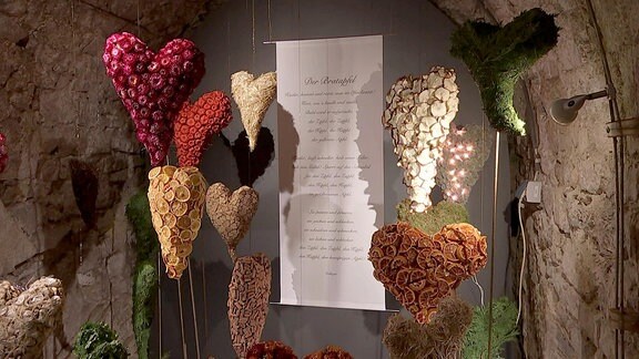 Herzen aus verschiedenen Materialien hängen neben einem Gedicht auf Papier von einer Decke.