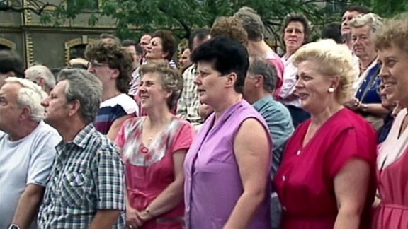 Eine große Gruppe Frauen in Kittelschürze und einige Männern auf einem Platz im Freien.