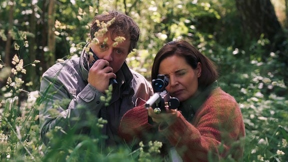 Ein Mann mit einem Mobiltelefon und eine Frau mit einem Gewehr im Anschlag in einem Waldstück.