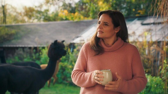Eine Frau im Pullover steht mit einer Tasse in den Händen in einem Tiergehege.