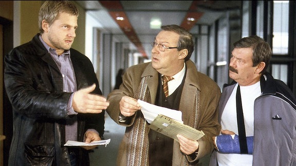 Die Kommissare Schmücke (Jaecki Schwarz, Mitte) und Schneider (Wolfgang Winkler, rechts) unterhalten sich mit einem Mann. Schmücke hält mehrere Akten in der Hand, Schneider trägt an einem Arm einen Gipsverband