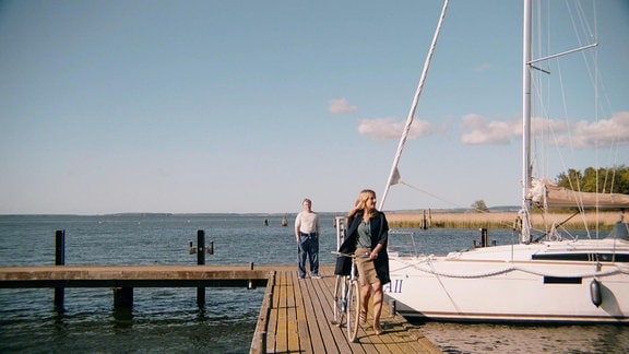 Eine Frau verlässt mit einem Fahrrad einen Bootssteg, auf dem ein Mann neben einem Segelboot steht.