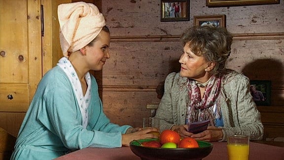 Eine junge Frau mit Handtuch um die Haare und eine ältere Frau sitzen an einem Tisch in rustikaler Stube.