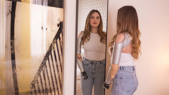 Eine junge Frau mit einem künstlichen Arm steht vor einem Spiegel.