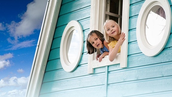 Ausschnitt aus einem Holzhausgiebeldach mit Türkisfarbenen Holz und 3 weißgerahmten Fenstern nebeneinander. Die beiden äußeren sind rund und aus dem mittleren eckigen schauen neugierig zwei ca. 4 bis 5 jährige Kinder. Hinter dem Dach ist blauer Himmel zu sehen.
