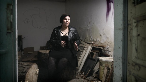 Eine schwarzhaarige Frau mit schwarzer Lederjacke in einem baufälligen Raum mit Fenstern, die am Bodeen stehen.