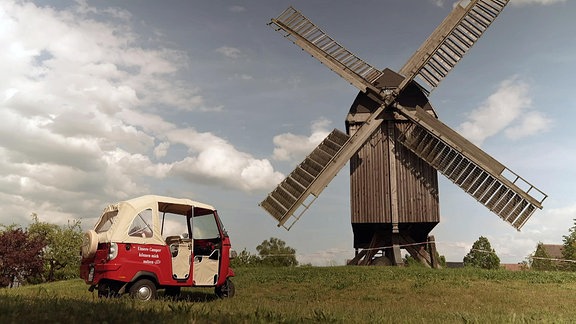 Ein rotes dreirädriges Fahrzeug mit weißem Verdeck steht am Fuße einer hölzernen Mühle auf einer Wiese.