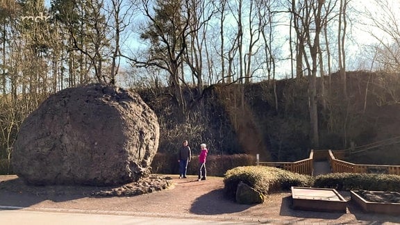 Zwei Personen stehen neben einem riesigen Lavastein in einem Park.