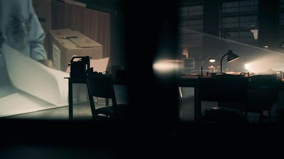 In einem dunklen Büro wird ein Schreibtisch beleuchtet, an der Wand läuft eine Videoprojektion mit Ermittlungsakten.