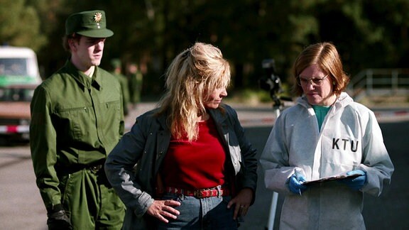Eine Frau in Zivil und eine Frau mit Schutzanzug, auf dem KTU steht, im Gespräch. Hinter ihnen steht ein junger Mann in einer grünen Uniform.