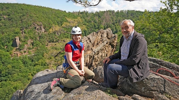 Ein Mann sitzt mit einer jungen Frau in Kletterausrüstung auf einem Felsen inmitten einer grünen Berglandschaft.