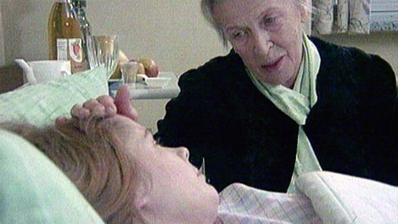 Ein Mädchen liegt im Krankenbett, ihre Oma sitzt daneben und streichelt ihre Stirn.