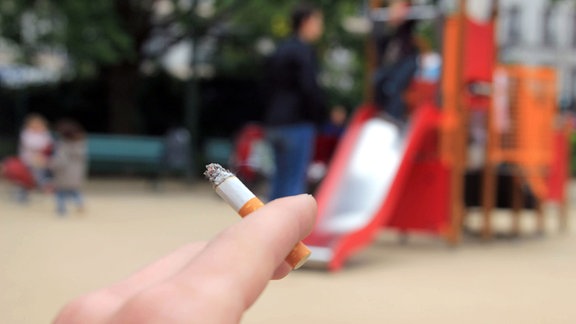 Symbolbild zum Rauchverbot auf Spielplätzen - im Vordergrund eine brennende Zigarette im Hintergrund ein Spielplatz.